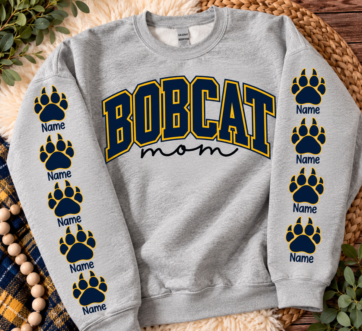This is A Digitally created Mock up of A heather gray crewneck sweatshirt laid flat on a woven mat with decorative greenery and a plaid blanket. The front has large arched 'BOBCAT' lettering outlined in yellow, with 'mom' in black script below.